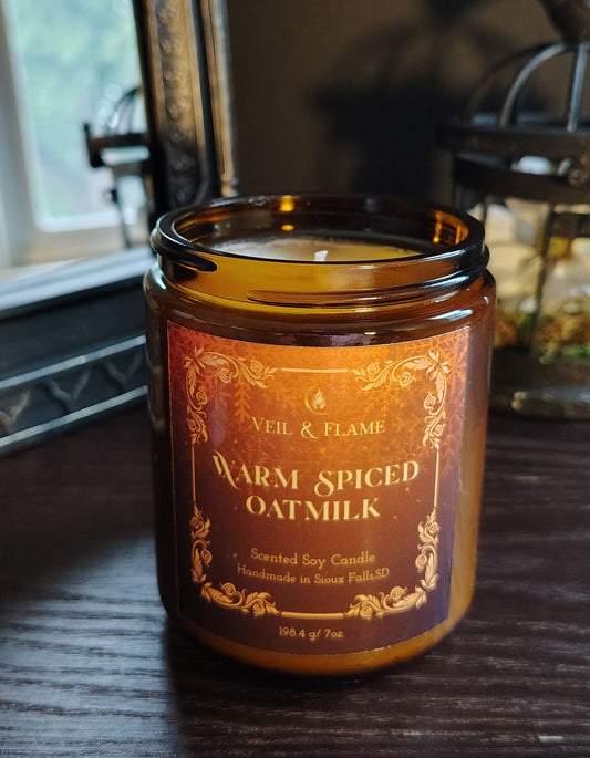 A warm amber glass jar candle labeled 'Veil & Flame Warm Spiced Oatmeal' sits on a dark wooden table in a cozy indoor setting with a mirror and lantern in the background.
