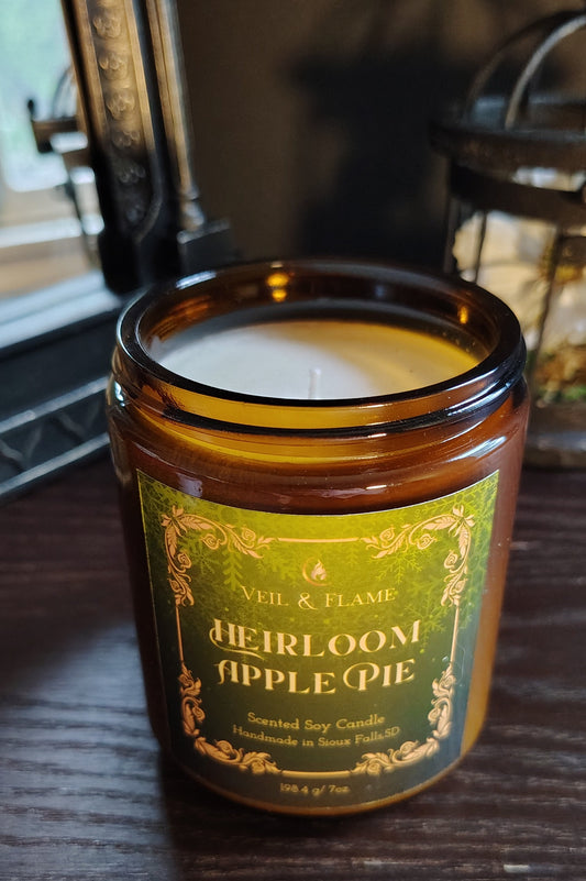 An amber glass jar candle labeled 'Heirloom Apple Pie' by Aile & Flame, sitting on a dark wooden table with a softly blurred indoor background.