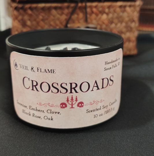 A black metal tin candle with a pink label reading 'CROSSROADS' and filled with white wax and decorated with dark wax flowers and herbs, set on a dark surface with a woven basket in the background.