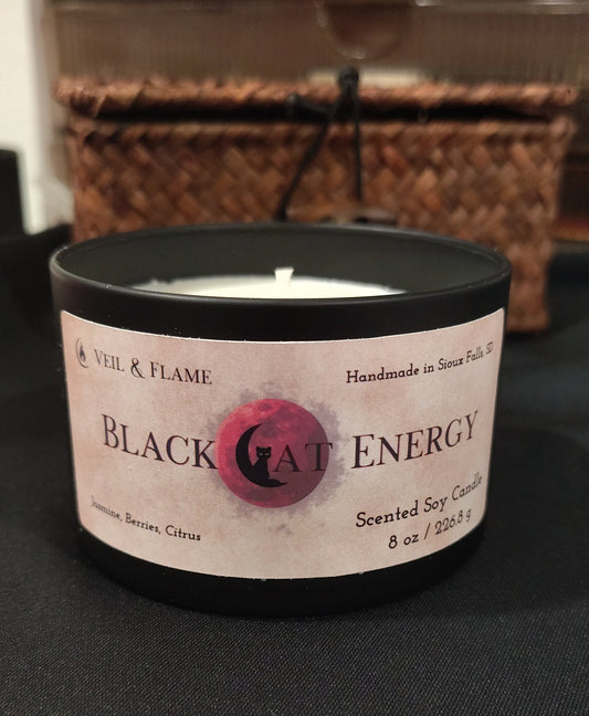 A black candle jar labeled 'Black Cat Energy' by Veil & Flame, placed on a dark surface with a woven basket in the blurred background.