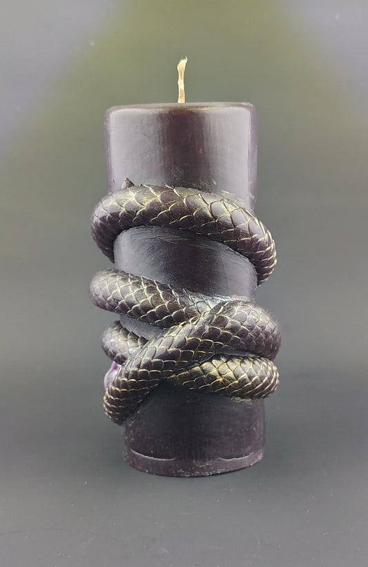 A dark purple pillar candle wrapped with a metallic gold snake sculpture, set against a dark gray background.