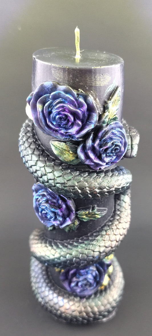 A black pillar candle wrapped with a metallic silver snake and decorated with three purple and blue roses with green leaves.