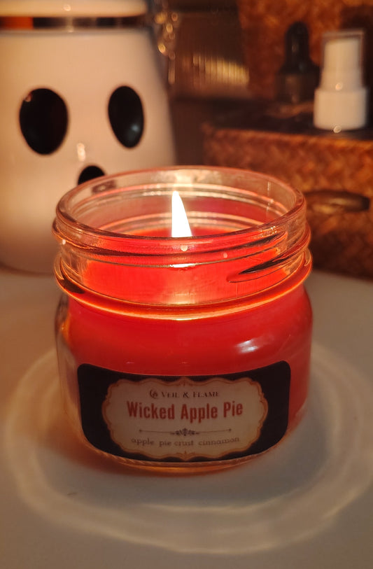 A lit red wax candle in a clear glass jar with a black label reading 'Wicked Apple Pie' by Veil & Flame, set on a white desk with a blurred desk background.