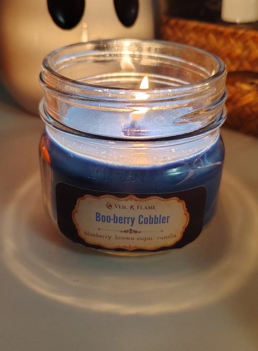 A lit blue candle in a clear glass jar with a black and cream label reading 'Veil & Flame Boo-berry Cobbler'.