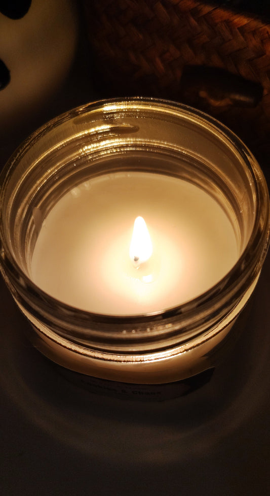 A lit jar candle with white wax in a clear glass container, photographed in warm, low lighting against a dark background.