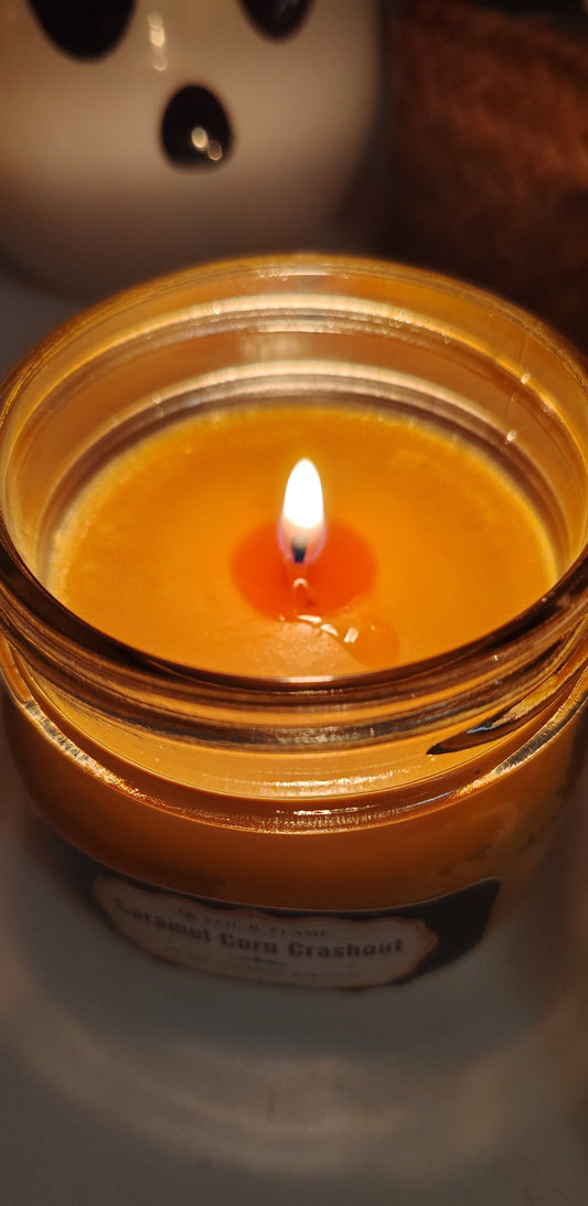 lit orange candle on white surface with ceramic figure and basket in the background. candle is titled Caramel Corn Crashout.