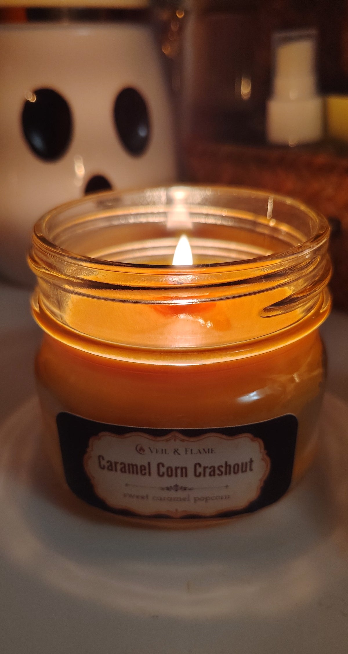 lit orange candle in clear glass jar titled Caramel Corn Crashout by Veil & Flame. Candle sits on white surface with a ceramic ghost in the background and a woven basket with small blurred items inside.
