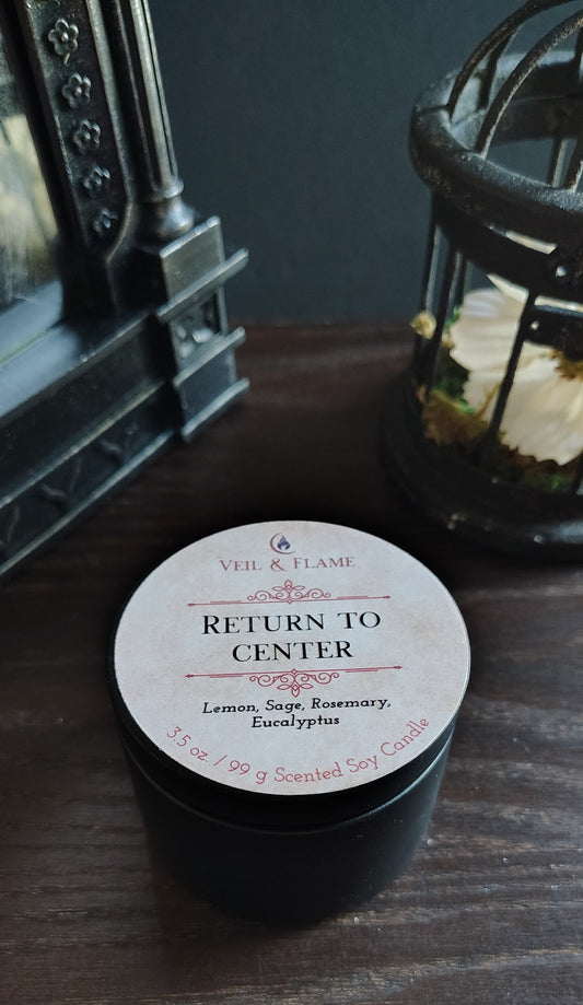 A black jar candle with a round white label reading 'RETURN TO CENTER' and 'Veil & Flame' at the top, placed on a dark wooden surface with decorative objects in the background.