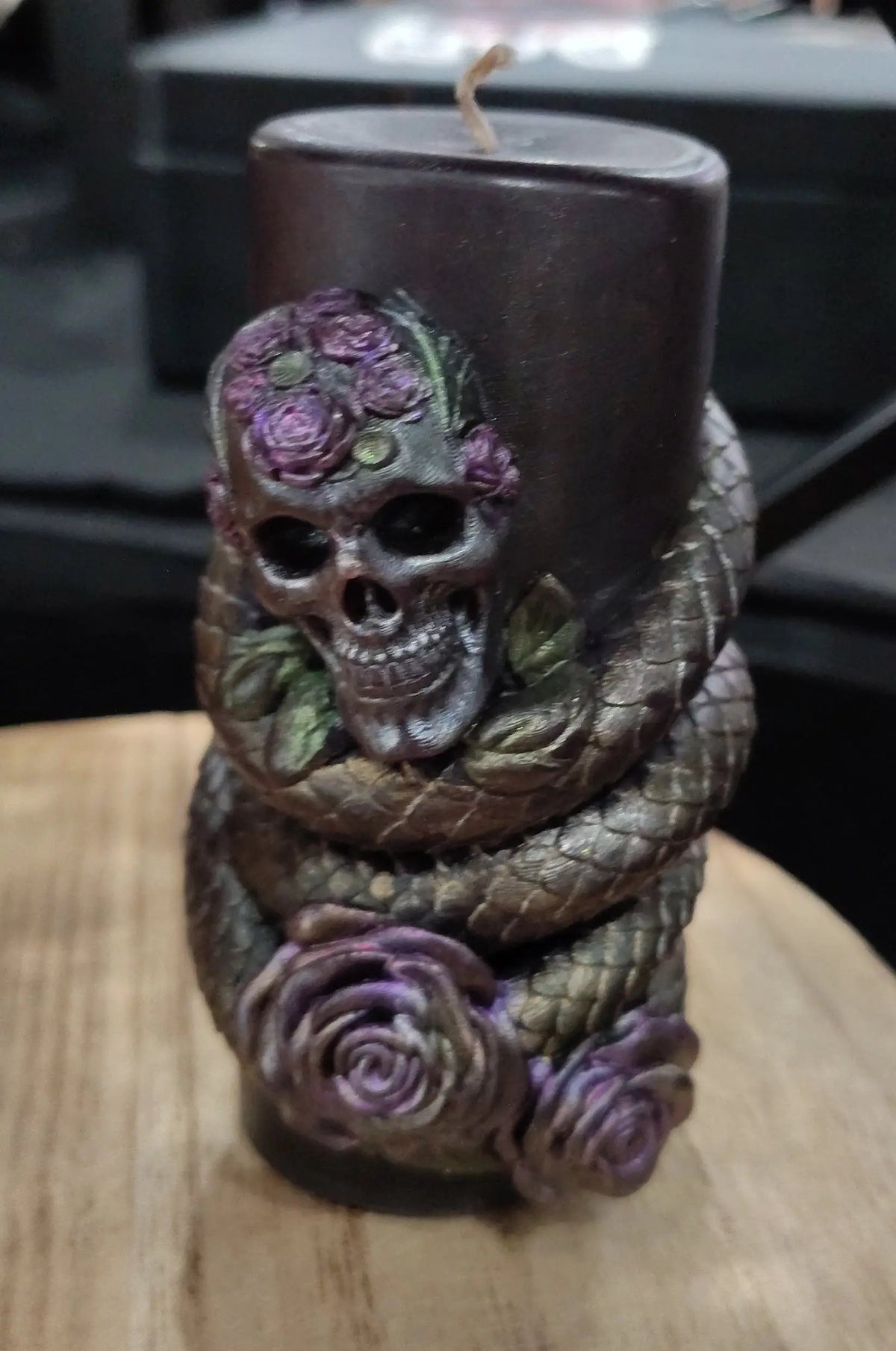 A black candle wrapped with a sculpted snake and decorated with purple roses and a skull on the front, displayed on a wooden surface.