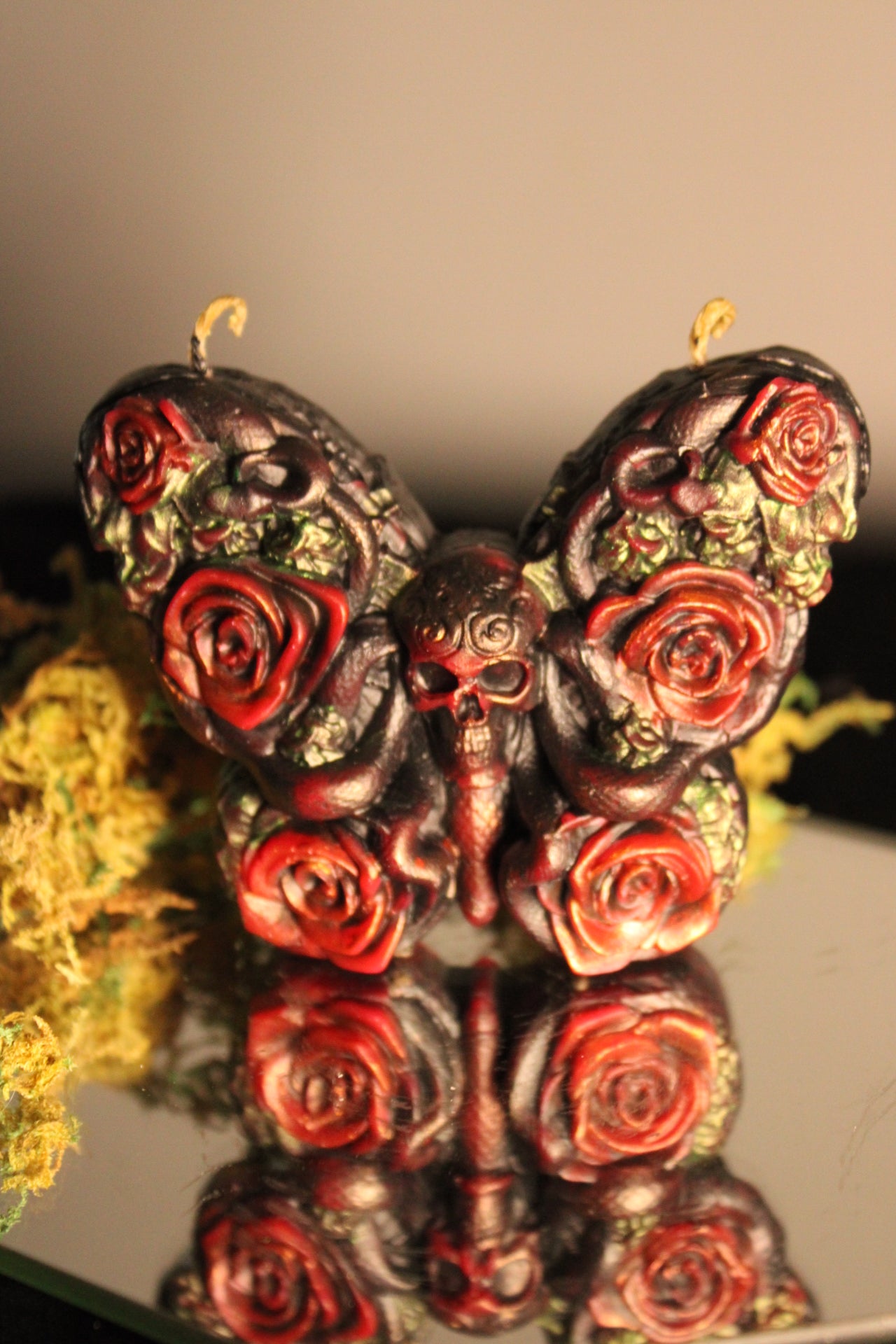 A decorative butterfly-shaped candle with a skull at the center and red roses across both wings, shown on a reflective surface with dried yellow flowers beside it.