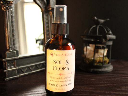 A bottle of Veil & Flame Sol & Flora room spray with a citrus and floral blend, placed on a dark wooden surface with a vintage mirror and a birdcage lantern in the background.
