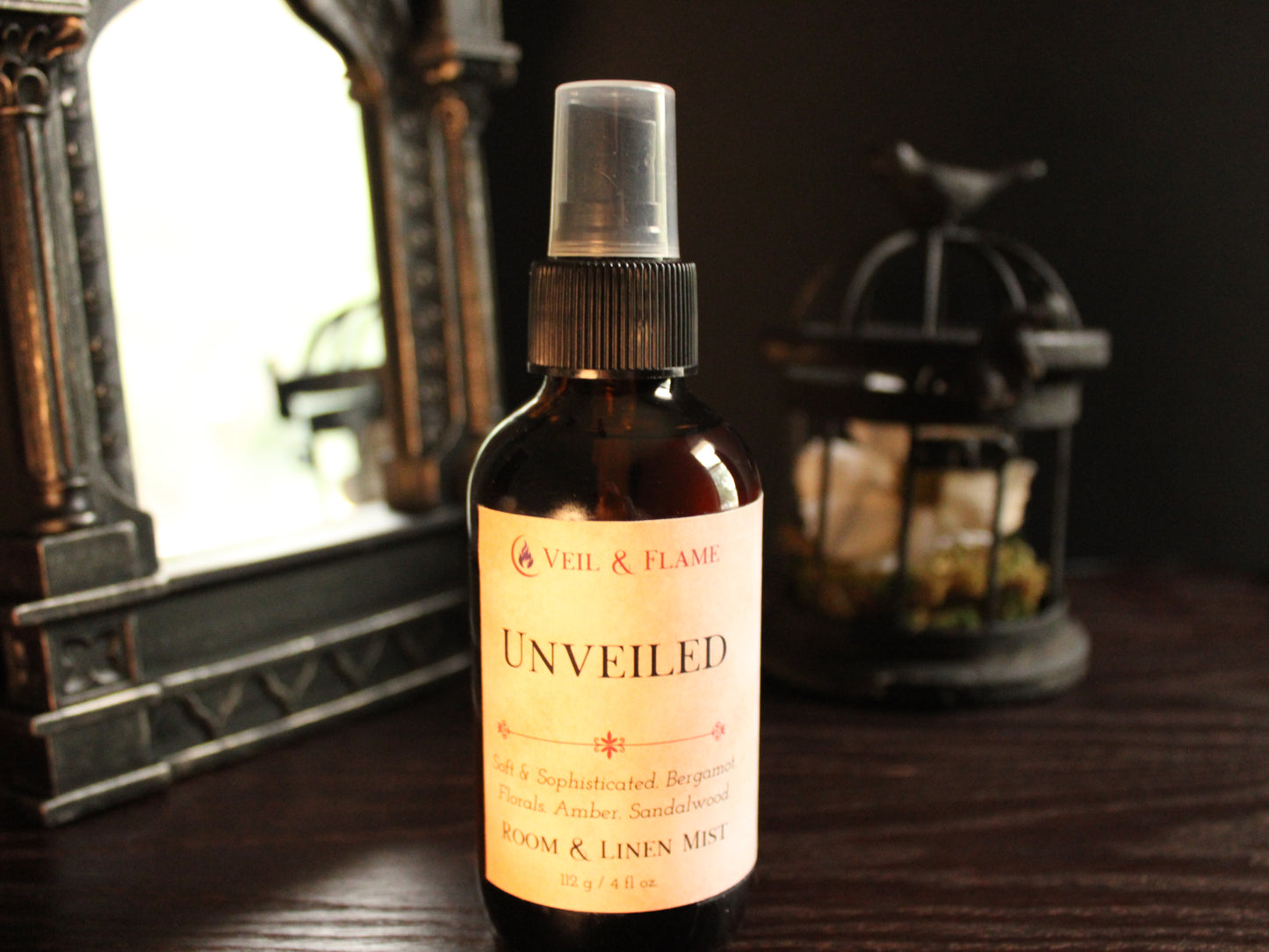 Amber spray bottle with cream labeling titled Unveiled. Label reads Soft & Sophisticated. Notes of bergamot, florals, amber, sandalwood. Bottle sits on wood surface with mirror and birdcage decoration blurred in the background. 
