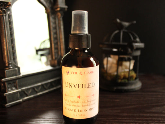 Amber spray bottle with cream labeling titled Unveiled. Label reads Soft & Sophisticated. Notes of bergamot, florals, amber, sandalwood. Bottle sits on wood surface with mirror and birdcage decoration blurred in the background. 