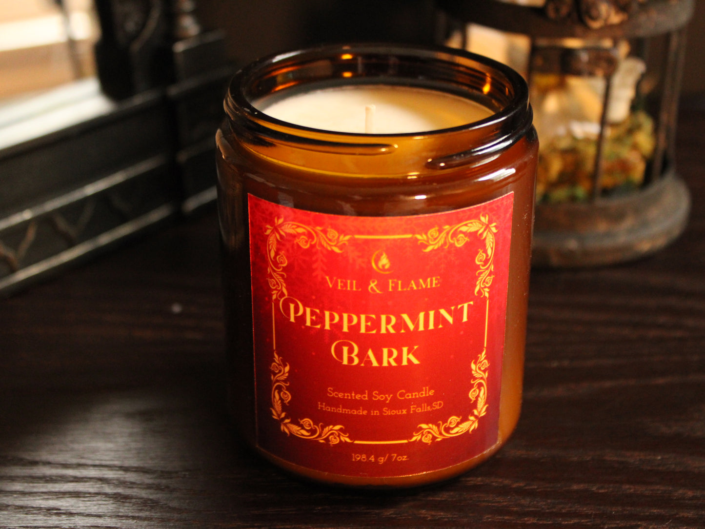 An amber glass jar candle labeled 'Peppermint Bark' by Veil & Flame, sitting on a dark wooden surface with a softly blurred background.