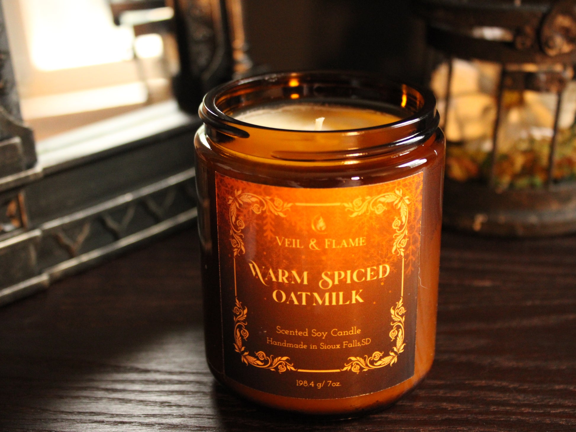 An amber glass jar candle labeled 'VEiL & FLAME Warm Spiced Oat Milk' sitting on a dark wooden table in a cozy indoor setting.