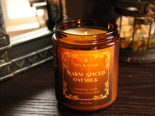 An amber glass jar candle labeled 'VEiL & FLAME Warm Spiced Oat Milk' sitting on a dark wooden table in a cozy indoor setting.