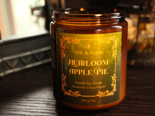 Amber jar candle with green and gold filigree label titled Heirloom Apple Pie by Veil & Flame. The candle sits on a dark wooden surface with a mirror and decor in the background. 