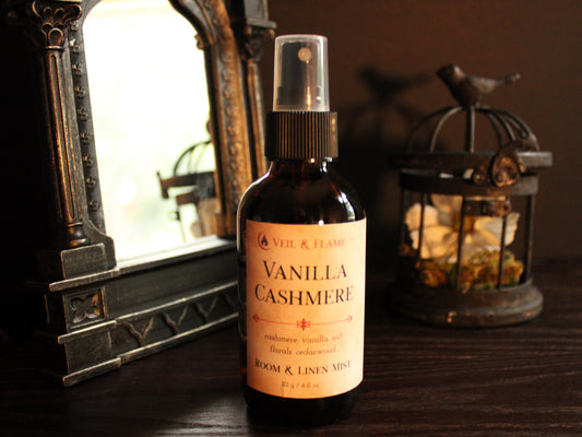 Amber bottle of room and linen spray called Vanilla Cashmere. The label is cream colored and lists scent notes of cashmere, vanilla, soft florals and cedarwood. The bottle sits on a wooden surface with a mirror and birdcage in the background. 