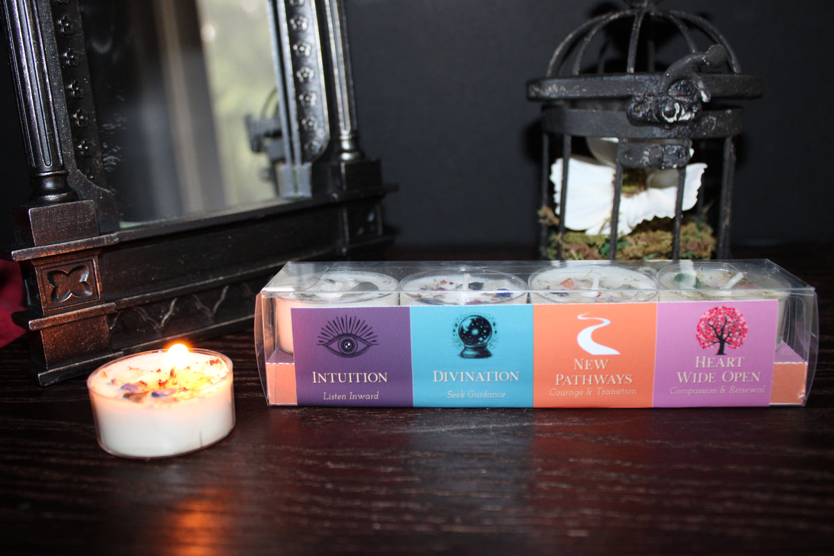 A boxed set of four tea light candles sits on a dark wooden table, with one lit candle in front and a mirror and decorative lantern in the background. The labels read Intuition (Listen Inward), Divination (Seek Guidance), New Pathways (Courage and Transition), and Flower Wild Open (Compassion and Renewal).