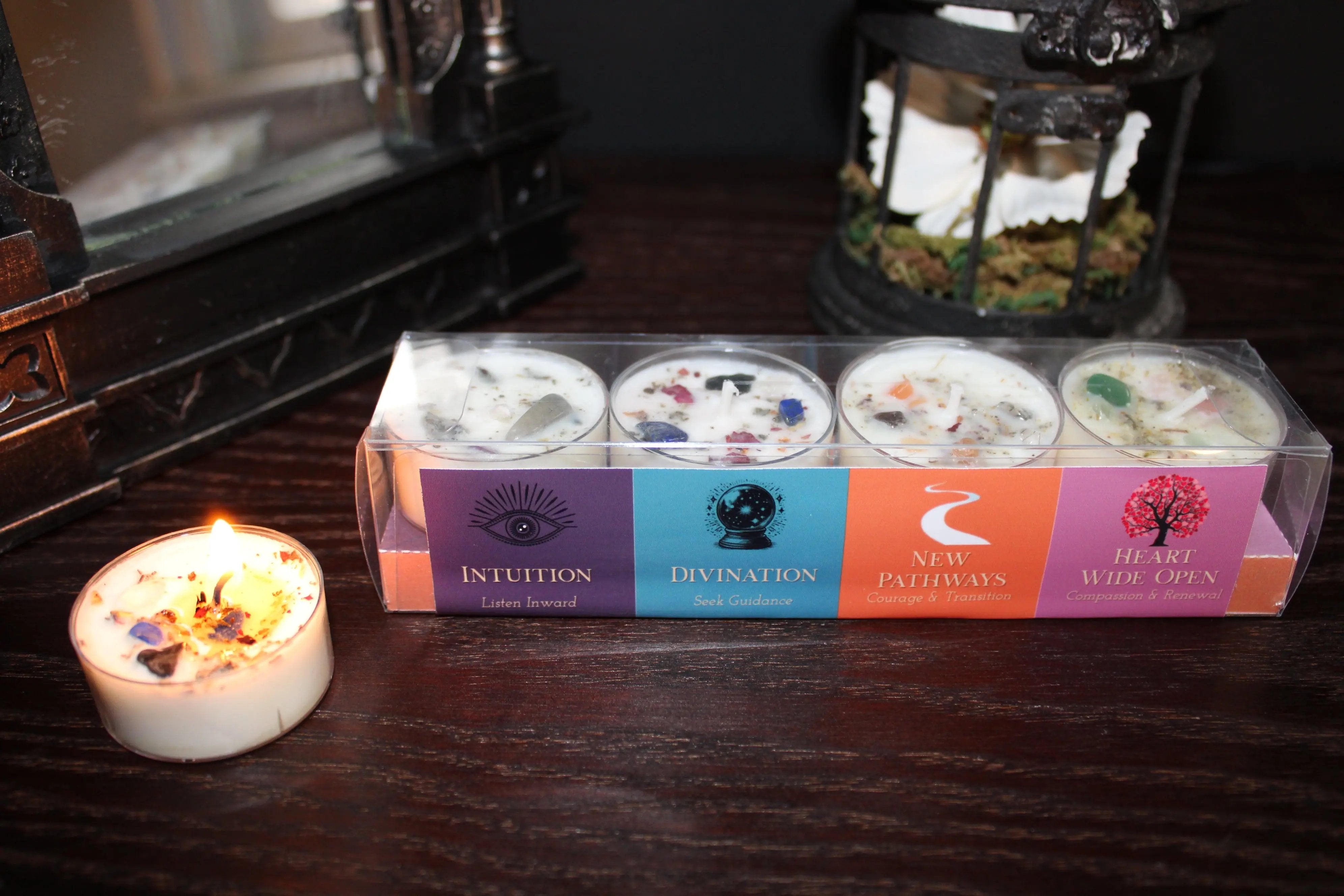 A boxed set of four small candles in a clear plastic case with colorful labels reading Intuition, Divination, New Pathways, and Wild Apps, placed on a dark wooden surface beside a lit tealight candle. Decorative lanterns and furniture are blurred in the background.