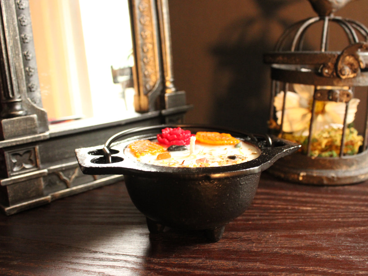 A black cast-iron cauldron-shaped candle decorated with colorful floral embellishments, sprinkled with a light dusting of herbs. A mirror and birdcage decor in background. Candle sits on wooden surface. 