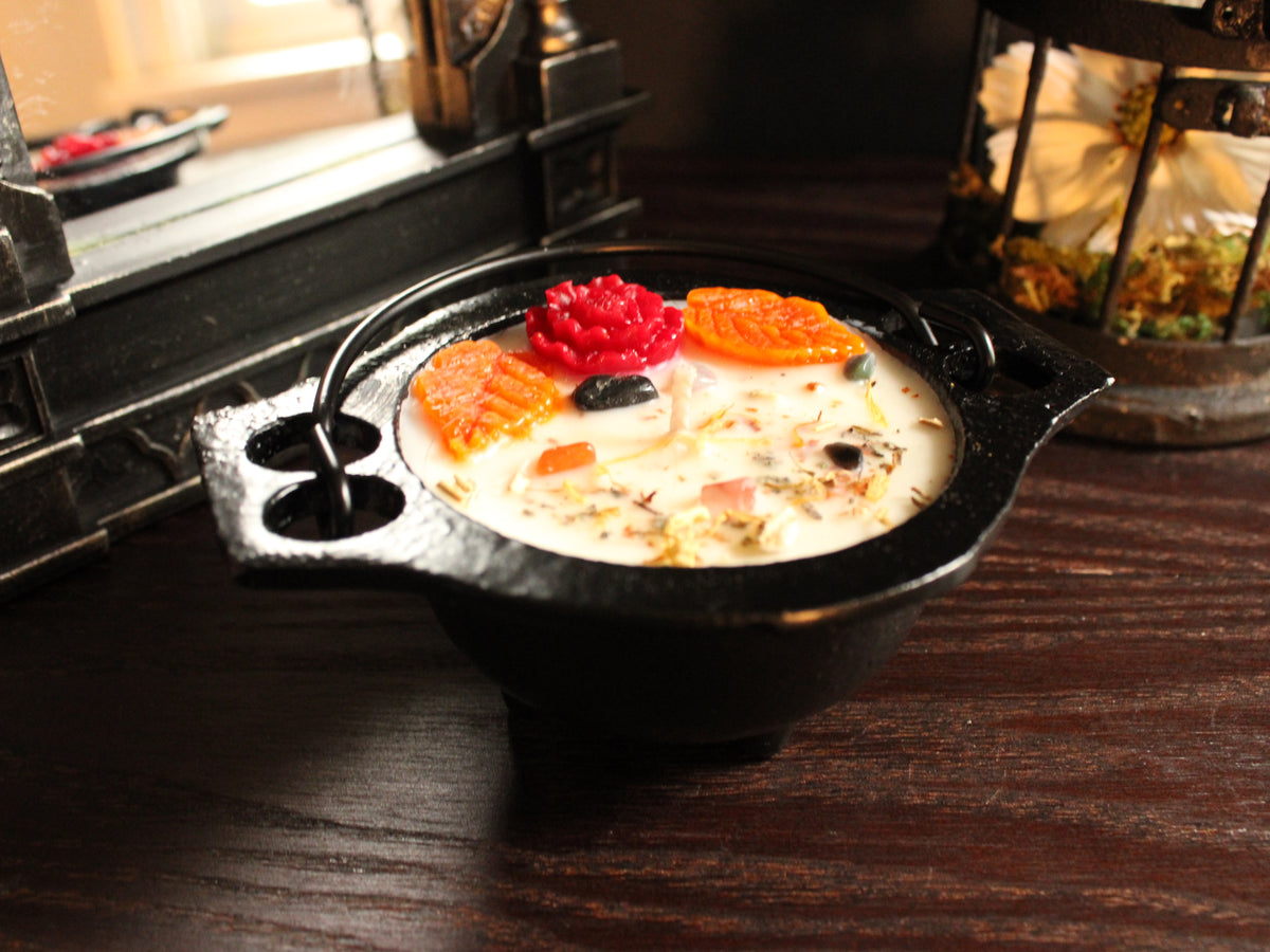 Small cauldron candle decorated with red and orange floral embellishments, sprinkled with a light dusting of herbs with mirror and birdcage decor in background. Candle sits on wooden surface. 