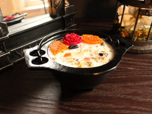 Small cauldron candle decorated with red and orange floral embellishments, sprinkled with a light dusting of herbs with mirror and birdcage decor in background. Candle sits on wooden surface. 