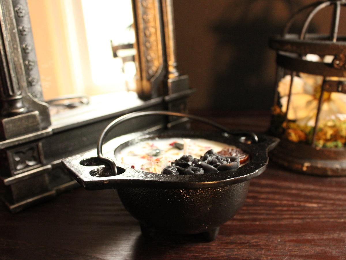 A black cast-iron cauldron-shaped candle decorated with black and brown floral embellishments, sprinkled with a light dusting of herbs. A mirror and birdcage decor in background. Candle sits on wooden surface. 