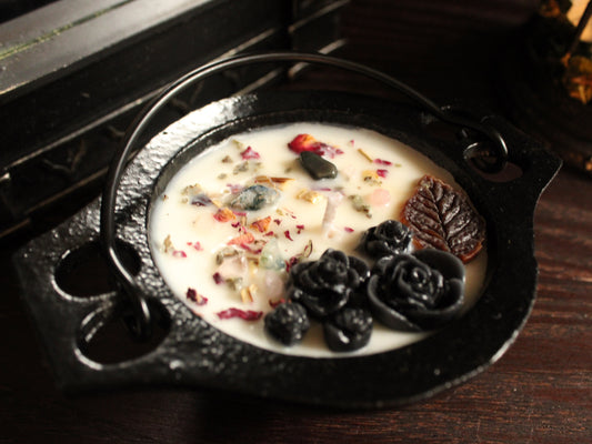 A black cast-iron cauldron-shaped candle decorated with black and brown floral embellishments, sprinkled with a light dusting of herbs. A mirror and birdcage decor in background. Candle sits on wooden surface. 