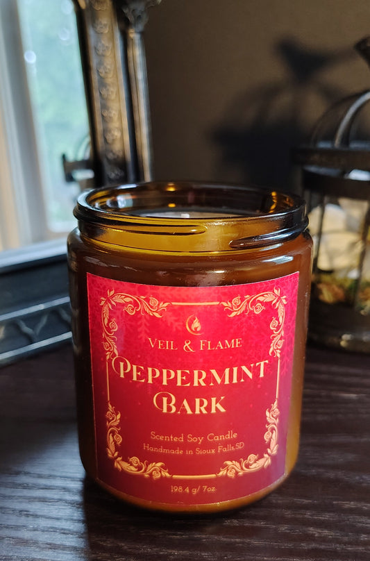 A scented soy candle in a amber glass jar with a red and gold label reading 'Vell & Flame Peppermint Bark,' sitting on a dark wooden table in an indoor setting.