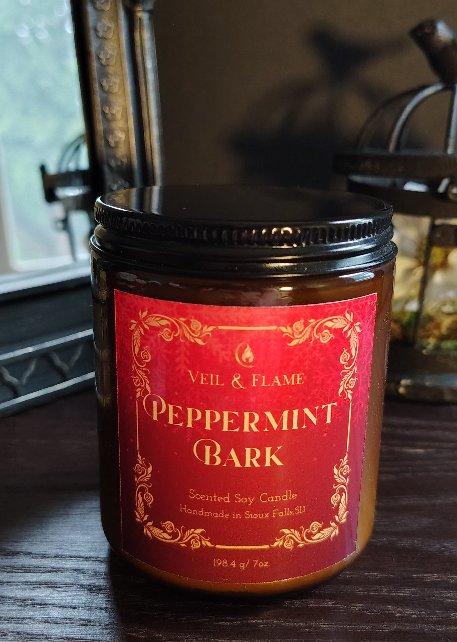 A jar candle labeled 'Veil & Flame Peppermint Bark' with a red label and black lid, sitting on a dark wooden table in a dimly lit room.