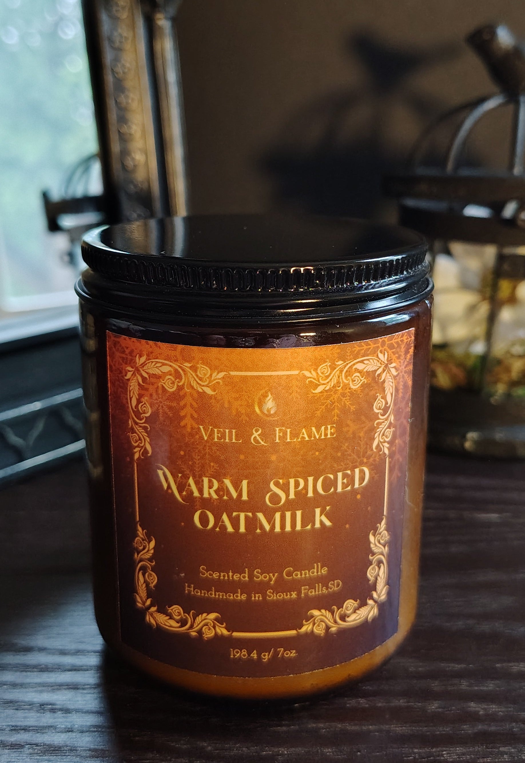 A jar candle labeled 'VEiL & FLAME Warm Spiced Oat Milk' sits on a dark wooden table in a dimly lit room with antique-style metal decor in the background.