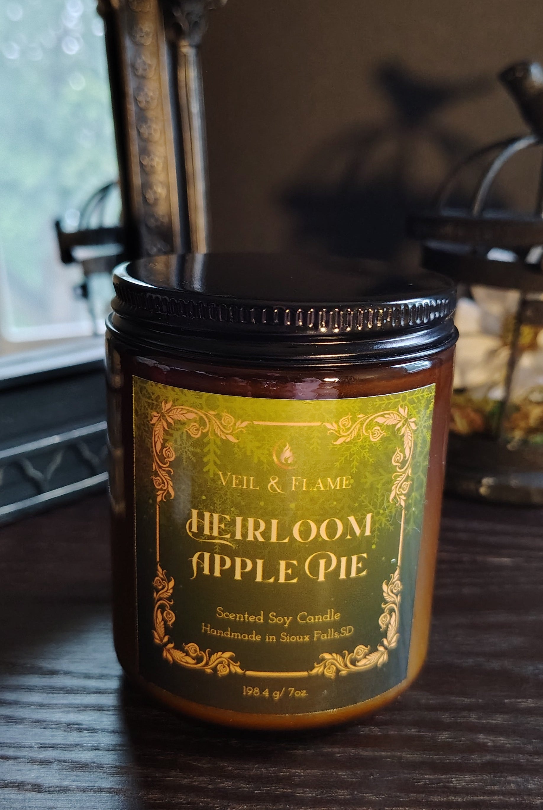 A jar candle labeled 'Heirloom Apple Pie' by Ash & Flame, sitting on a dark wooden table in a dimly lit room.