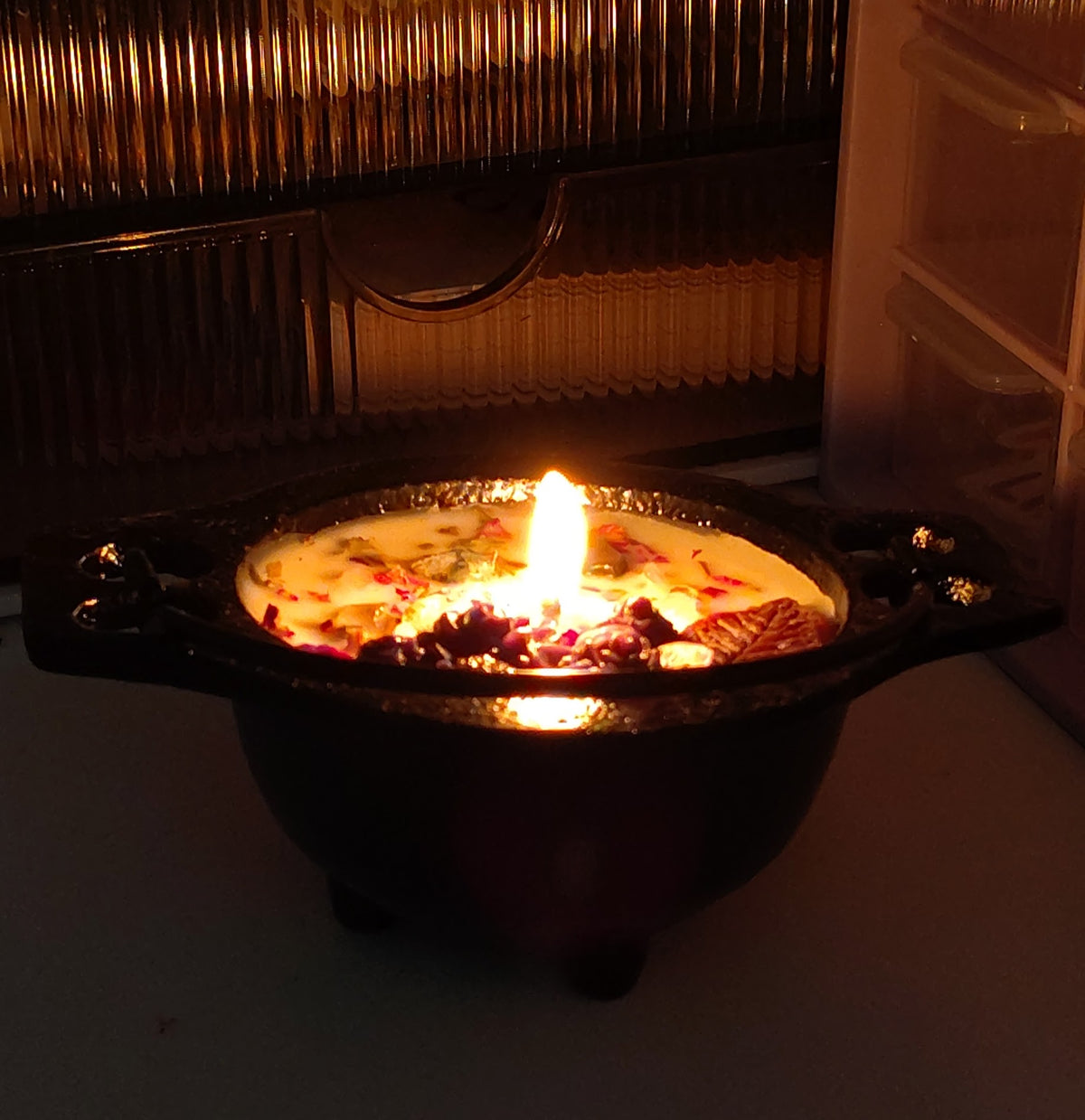 A dark cast-iron pot candle with a single lit wick, filled with wax and embedded botanical elements, set against a dark, warm-toned background.