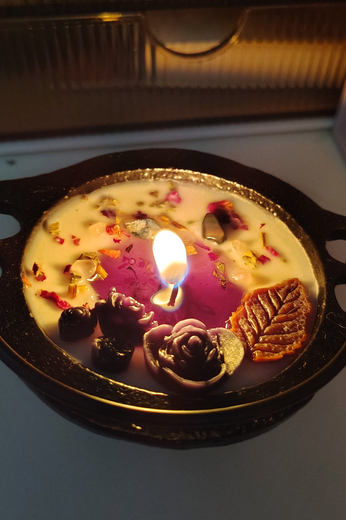 A decorative candle in a small dark cauldron, lit with a small flame. The wax is creamy off-white with embedded dried flowers, petals, and a leaf. Shows the meltpool of this candle being a purple color in center. 