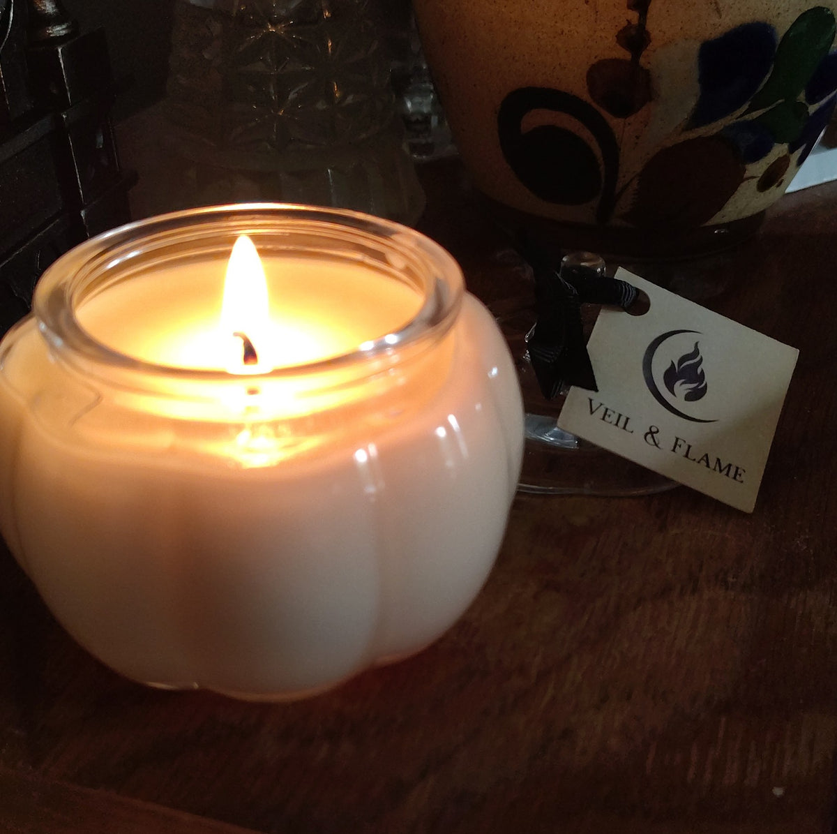 A lit cream-colored candle in a ribbed glass jar sits on a wooden surface next to a black ribbon and a paper tag reading 'VEIL & FLAME'.
