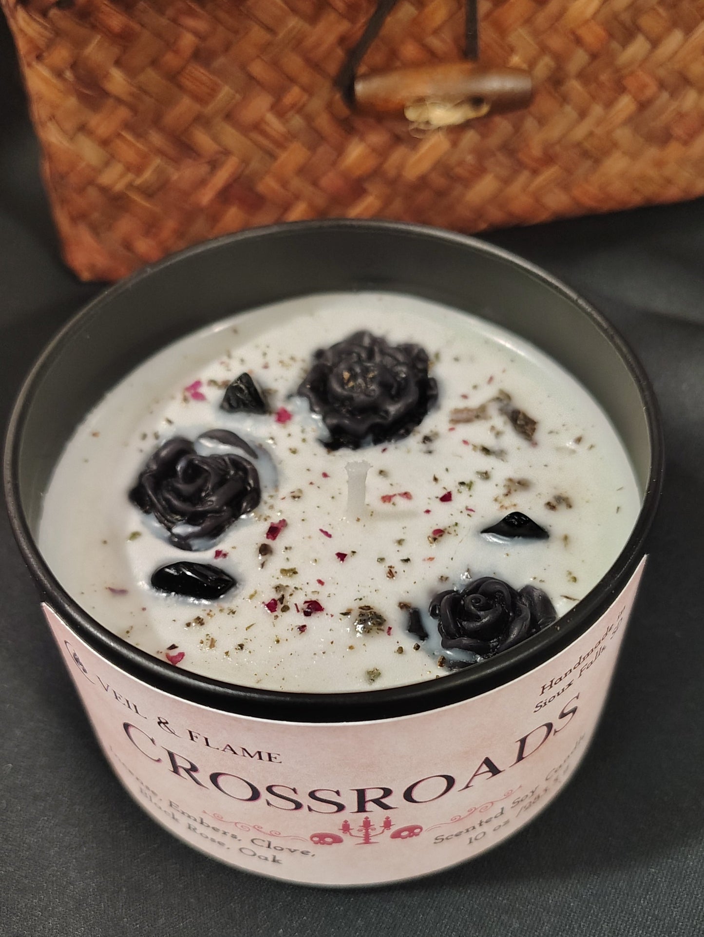 A black metal tin candle with a pink label reading 'CROSSROADS' and filled with white wax and decorated with dark wax flowers and herbs, set on a dark surface with a woven basket in the background.