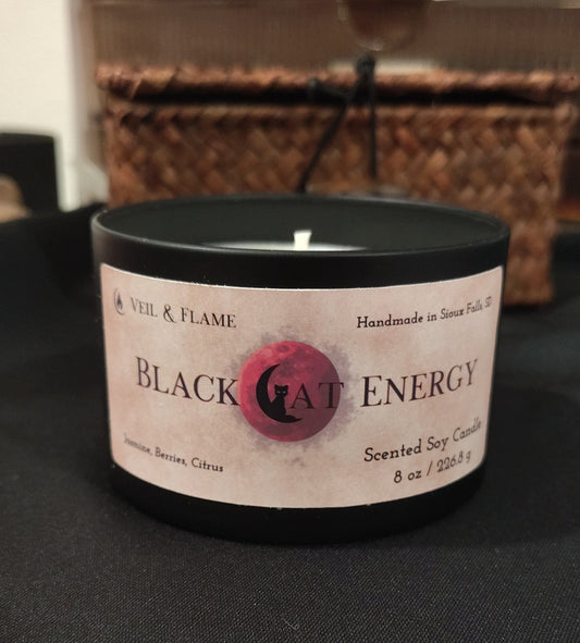 A black glass jar candle with a white label reading 'BLACK CAT ENERGY' and a pink cat silhouette logo, placed on a dark surface with a woven basket in the background.