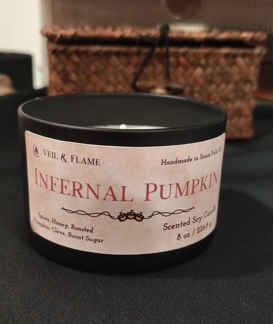 A black candle tin labeled 'Infernal Pumpkin' by Veil & Flame, placed on a dark surface with a blurred home background.