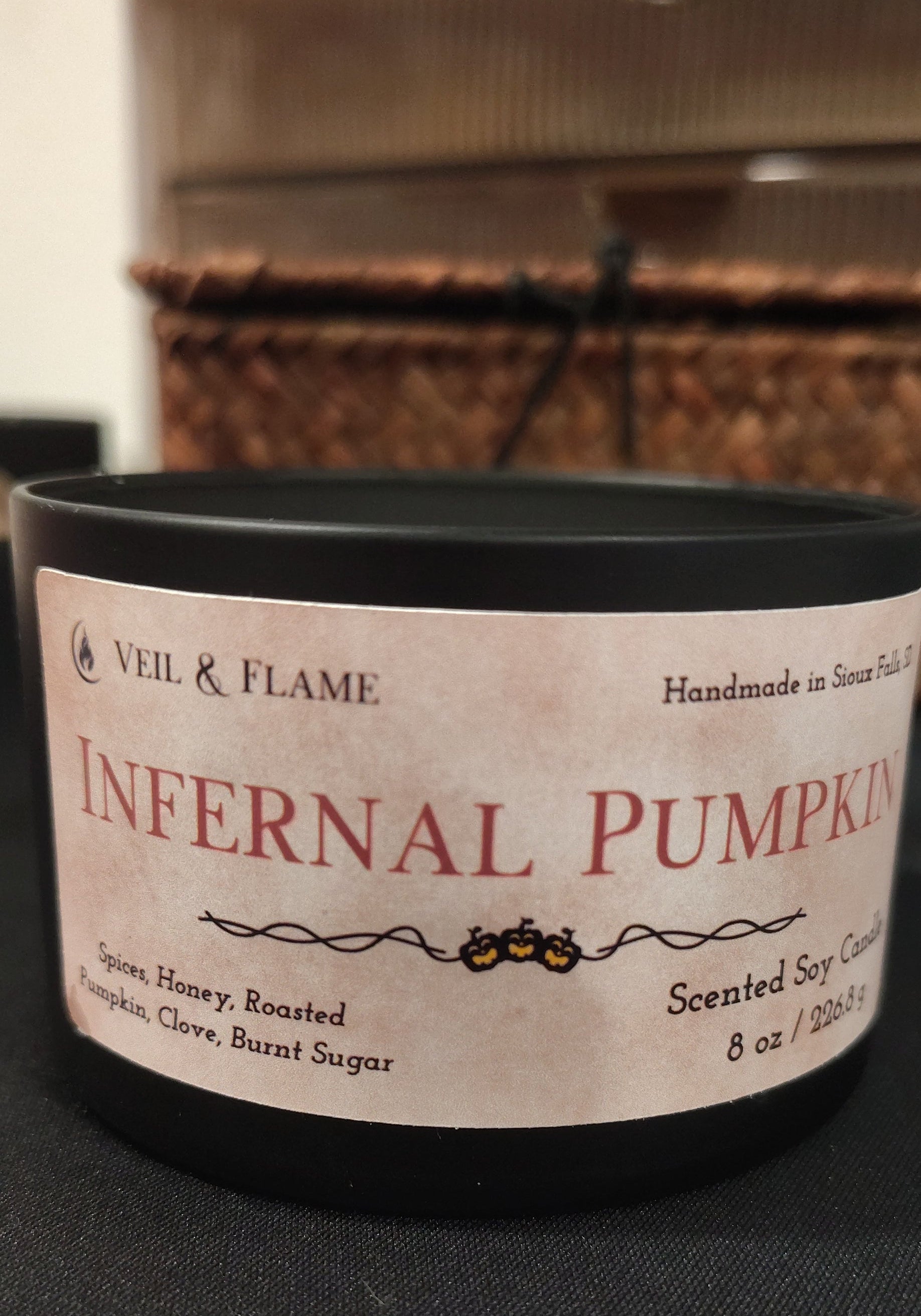 A black scented soy candle tin with a cream-colored label reading 'Infernal Pumpkin' by Veil & Flame, photographed on a dark surface with a blurred indoor background.