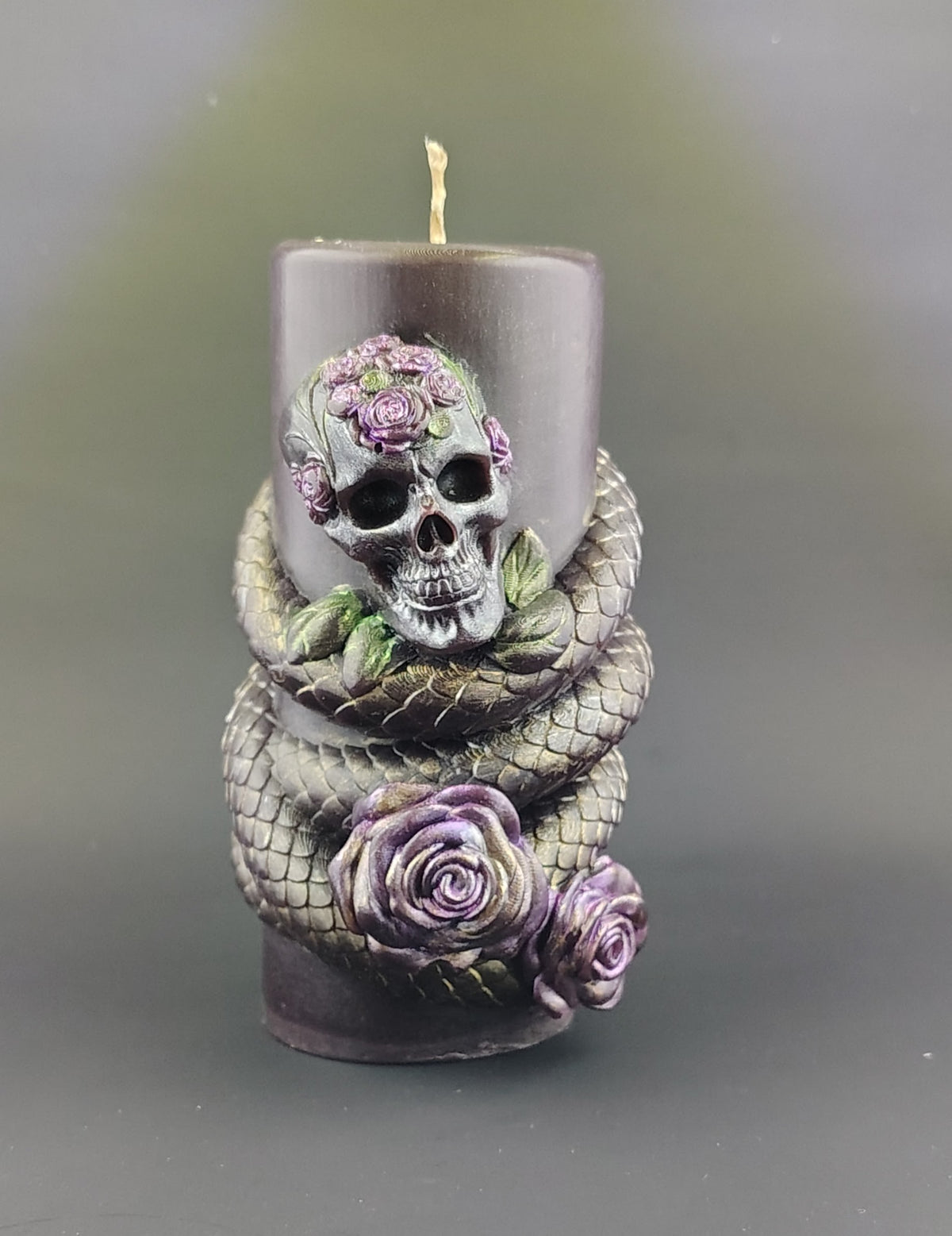 A dark red pillar candle featuring a sculpted skull adorned with purple roses and a gray snake wrapped around the base, set against a soft gray background.