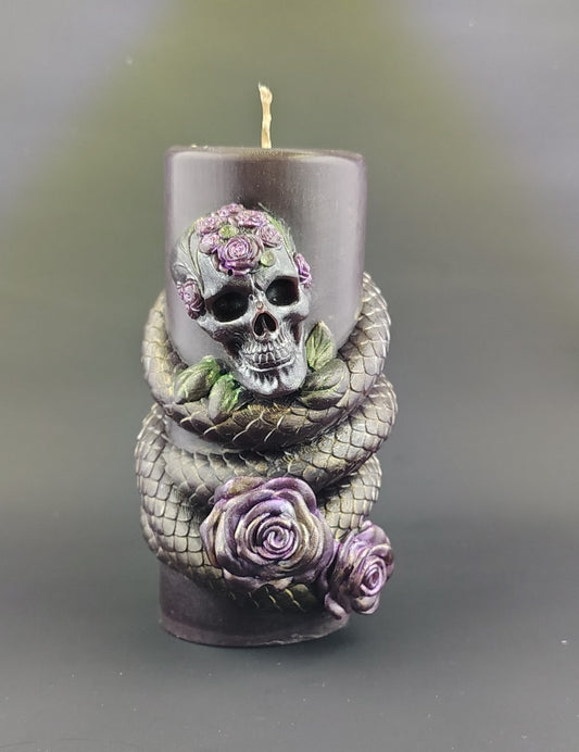A dark red pillar candle featuring a sculpted skull adorned with purple roses and a gray snake wrapped around the base, set against a soft gray background.