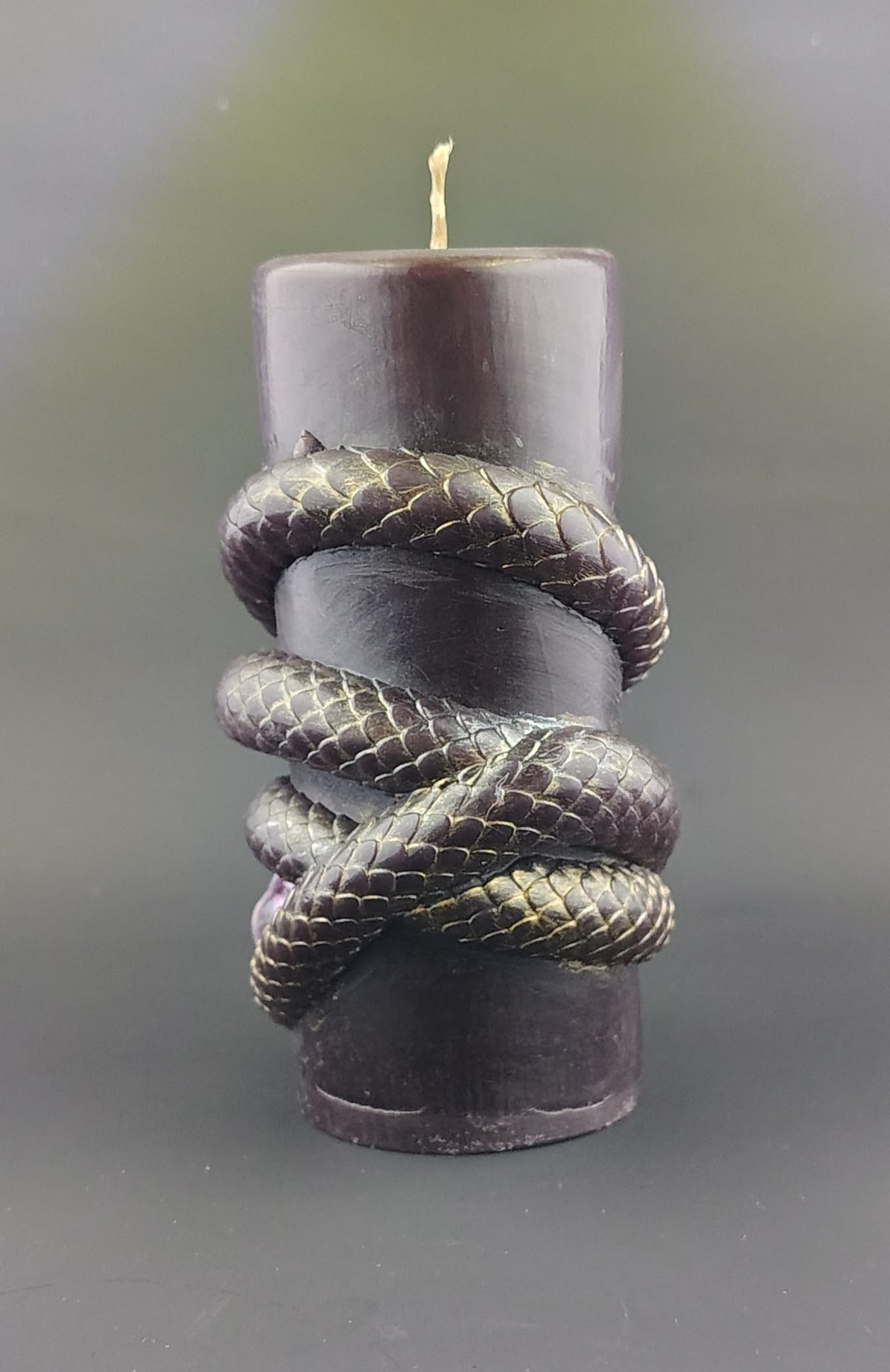 A dark purple pillar candle wrapped with a metallic gold snake sculpture, set against a dark gray background.