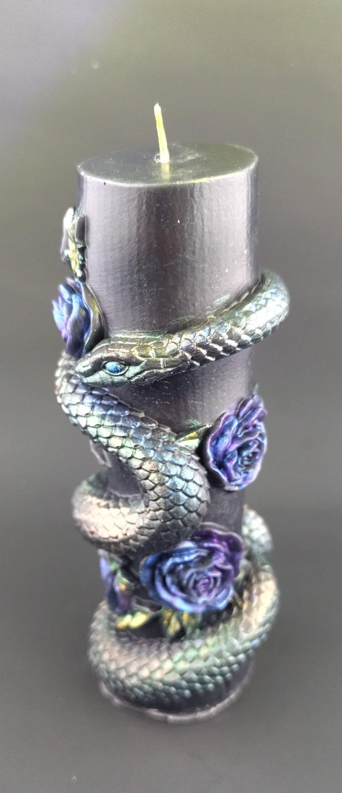 A black pillar candle wrapped with a metallic iridescent snake and decorated with blue-purple roses, set against a dark gray background.
