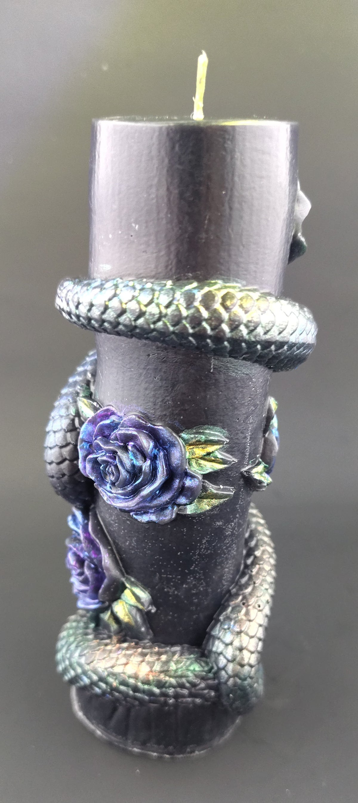 A black pillar candle wrapped with a metallic iridescent snake and decorated with purple roses, shown against a dark gray background.