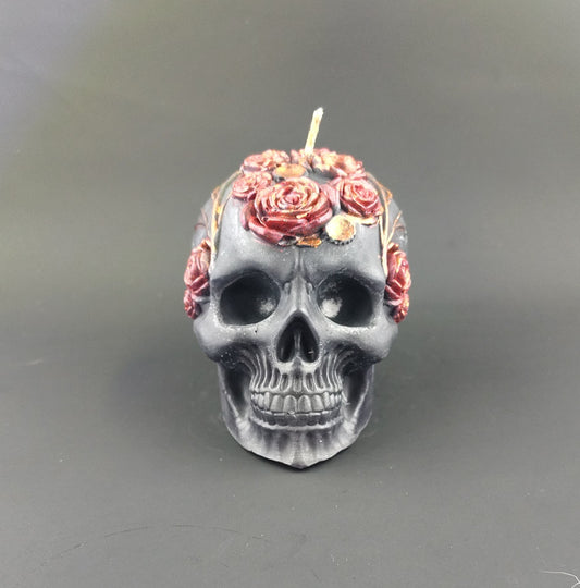 A skull-shaped candle in dark gray with red and pink roses on top and along the sides, photographed against a dark gray background.