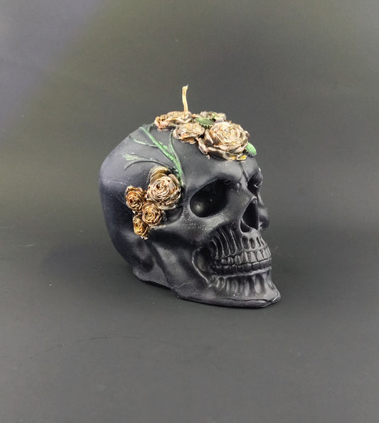 A black skull-shaped candle decorated with gold-toned dried roses and green leaves, set against a dark gray background.