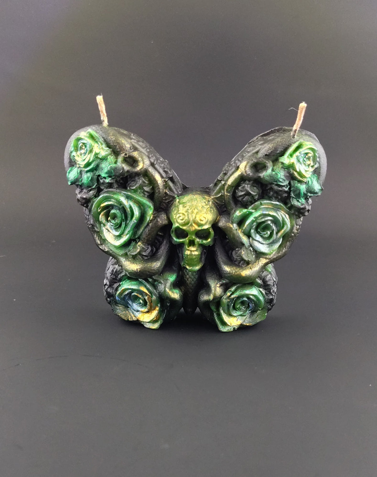 Butterfly shaped candle in black with green accented florals and a skull for a head. Background is a plain black studio shot. 