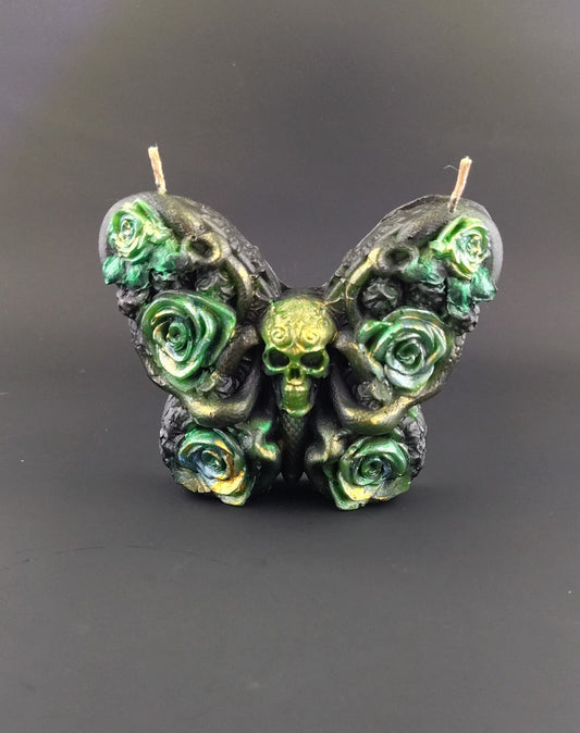 Butterfly shaped candle in black with green accented florals and a skull for a head. Background is a plain black studio shot. 