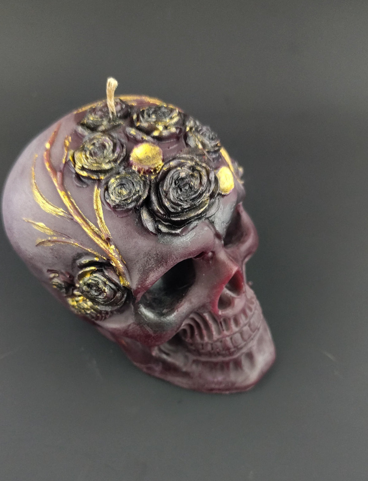A decorative skull-shaped candle in a dark purple hue, embellished with black and gold rose details on top, photographed on a dark gray background.